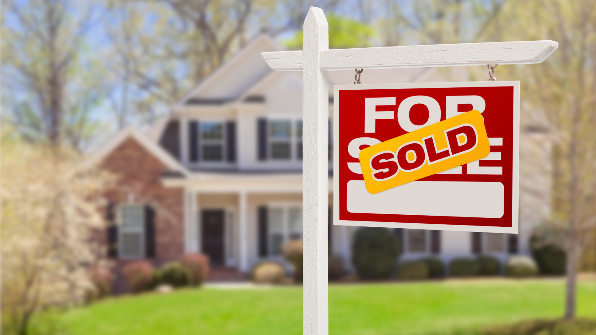 A real estate “For Sale” sign with a “Sold” sticker in front of a landscaped home